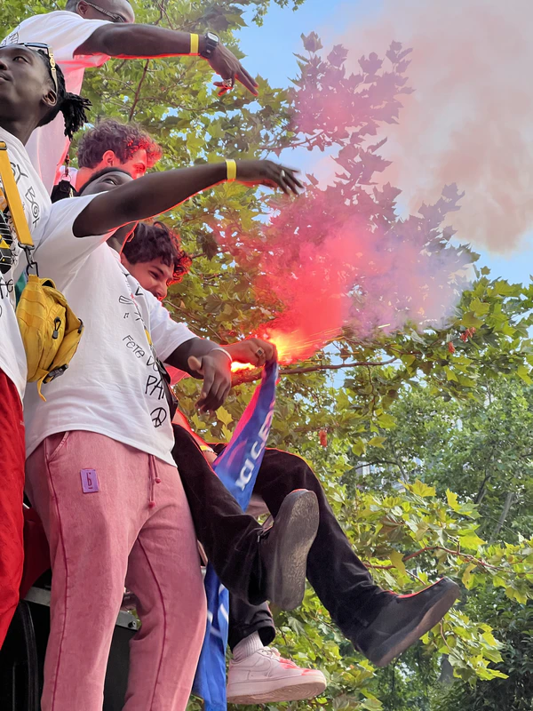 Crowd celebrating under trees, one fan holding a lit red smoke flare overhead